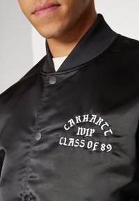 Homme portant une veste varsity en satin noir avec col côtelé noir et texte brodé blanc "Carhartt WIP Class of 89" sur la poitrine.