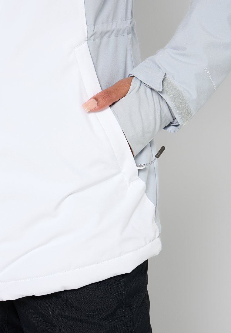 Hand in einer hellgrauen Jackentasche mit Klettverschluss, der Körper der Jacke ist weiß und schwarze Hosen sind sichtbar.
