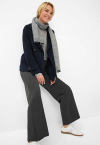 Marineblauer Doppelreiher-Mantel, grauer Strickschal, hellgrauer Pullover, graue weit geschnittene Hose und weiße Sneakers mit brauner Sohle.