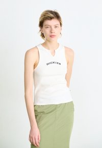 Haut sans manches en ribbed blanc avec un col rond et un détail en découpe, arborant le texte noir "DICKIES" sur la poitrine, associé à un pantalon vert.