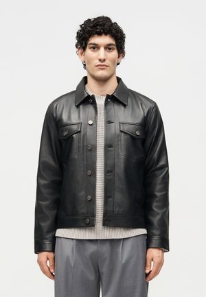 Jeune homme aux cheveux courts et bouclés portant une veste en cuir noire boutonnée, un pull léger en tricot et un pantalon gris, debout devant un fond blanc.