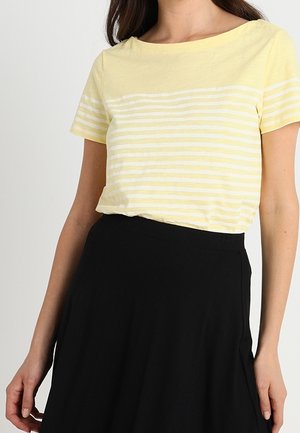 Femme portant un haut à manches courtes rayé jaune clair et blanc, rentré dans une jupe noire taille haute, sur un fond neutre.