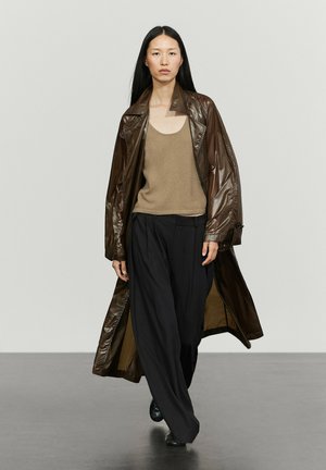 Femme aux longs cheveux noirs porte un trench-coat en cuir marron, un débardeur beige, un pantalon noir ample et des chaussures noires en marchant vers l'avant sur un sol gris.