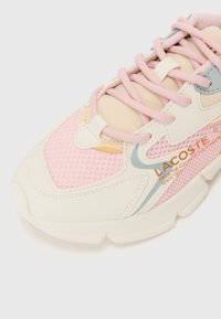 Lacoste-sko med mesh-stoff i lys rosa og kremfarge, med blå detaljer, flate snøringer og en teksturert såle.