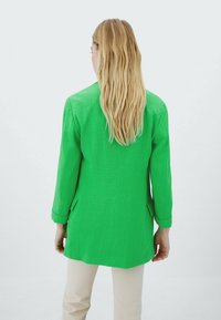 Personne aux cheveux longs et blonds portant un blazer vert vif et un pantalon beige, debout de dos sur un fond blanc uni.