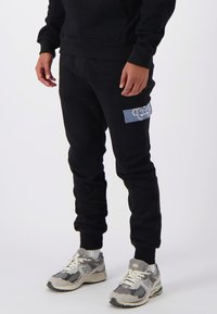 Zwarte joggingbroek met een zijvak en een blauwe patch. Gemaakt van een zachte stof, hebben ze een getailleerde pasvorm en ribbels aan de onderkant.