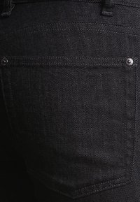 Jeans de mezclilla negra con una superficie texturizada, que presentan un bolsillo trasero y piezas de botón metálico, mostrando un diseño ajustado y ceñido.