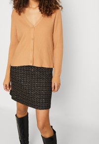 Cárdigan beige de canalé con escote en V y cinco botones, combinado con una falda mini negra texturizada con detalles en dorado, llevado con botas altas negras.