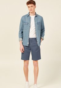 Jeansskjorta i ljusblått med knappframsida och bröstfickor, kombinerad med marinblå shorts. Modellen bär en vit t-shirt och sneakers.