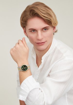 Giovane uomo con camicia bianca che indossa un orologio da polso in metallo bicolore con quadrante verde, che guarda leggermente di lato contro uno sfondo chiaro e semplice.