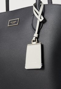 Borsa a tote in pelle nera testurizzata con doppi manici, caratterizzata da un'etichetta bianca con un logo in oro stampato. Le rifiniture includono hardware dorato.
