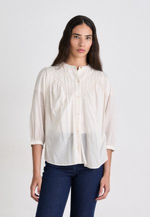 Camicia button-up di un beige chiaro con righe verticali, maniche corte e colletto alla coreana. Tessuto testurizzato con sottili dettagli ricamati.