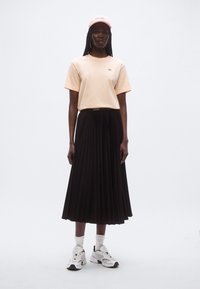 T-shirt pêche clair avec un petit logo, associé à une jupe plissée noire. Stylisé avec une casquette rose et des chaussures de sport blanches. Design texturé, décontracté.