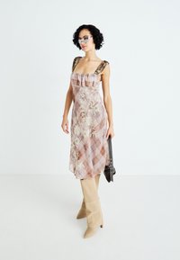Abito in tessuto trasparente con motivo floreale e a quadretti, con scollo arruffato e spalline marroni. La modella indossa stivali beige fino al ginocchio e occhiali da sole.