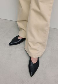 Zwarte leren schoenen met spitse neuzen en een uitsnijd ontwerp. Draagt een lichtbeige wijde broek en staat op een grijze vloer.