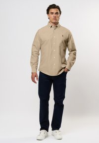 Uomo in piedi che indossa una camicia beige con bottoni, jeans blu scuro e scarpe bianche su uno sfondo chiaro e semplice.