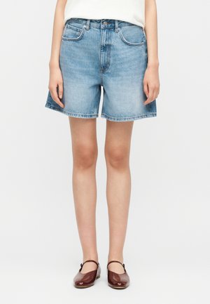 Mujer vistiendo shorts de mezclilla azul claro, camiseta blanca y zapatos Mary Jane marrones, de pie contra un fondo blanco liso.