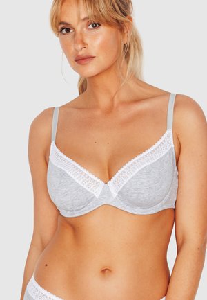 Donna bionda dalla pelle chiara che indossa un reggiseno grigio con rifiniture in pizzo bianco e spalline regolabili, su uno sfondo chiaro e uniforme.