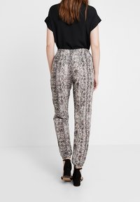 Kvinna som bär en svart topp och byxor med hög midja i ormskinnsmönster med elastiska manschetter, kombinerat med svarta öppna blockklackar, som står mot en vit bakgrund.