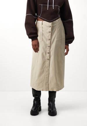 Femme portant une jupe midi beige à boutons sur le devant avec de grandes poches, des bottes noires épaisses, et un sweat à capuche court marron foncé avec des cordons blancs.