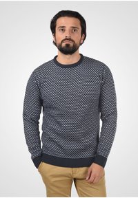 Mørkeblå sweater med et tekstureret mønster af små hvide former, ribbede manchetter og kant, rund halsudskæring, båret med khakifarvede bukser.