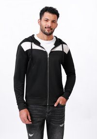 Haut à capuche zipé noir avec accents blancs sur les épaules, fabriqué en tissu doux, offrant une coupe décontractée, des poignets à côtes et des poches avant.