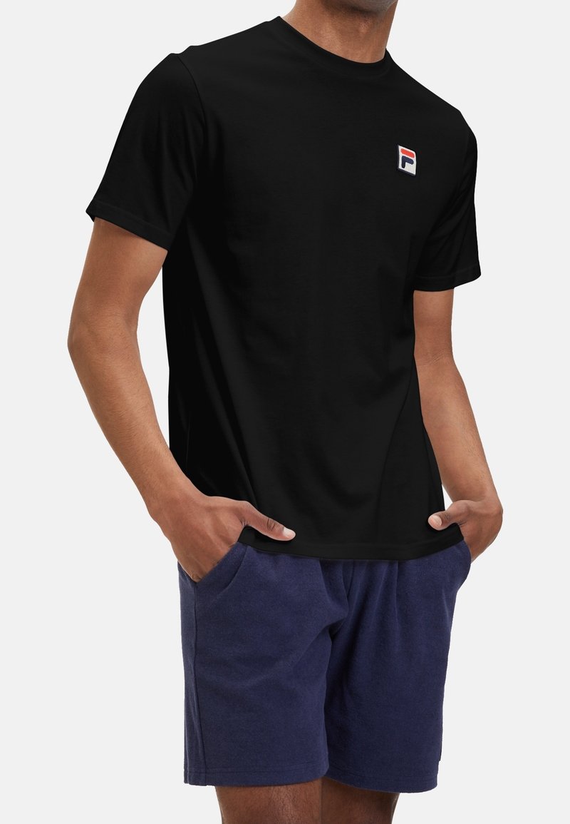 Black cotton t-shirt with crew neck, featuring a small colorful logo on the left chest. Model wears navy shorts with pockets.