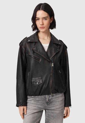 Nuori nainen, jolla on musta nahkainen moottoripyörätakki valkoisen topin ja harmaiden farkkujen päällä, yksinkertaista vaaleaa taustaa vasten.