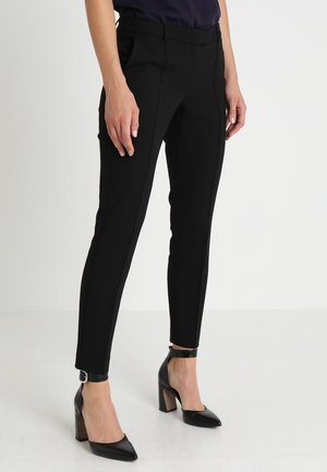 Nainen, joka pukeutuu kapeisiin mustiin housuihin ja mustiin korkokenkiin nilkkahihnoilla, seisoo yksinkertaisen valkoisen taustan edessä.