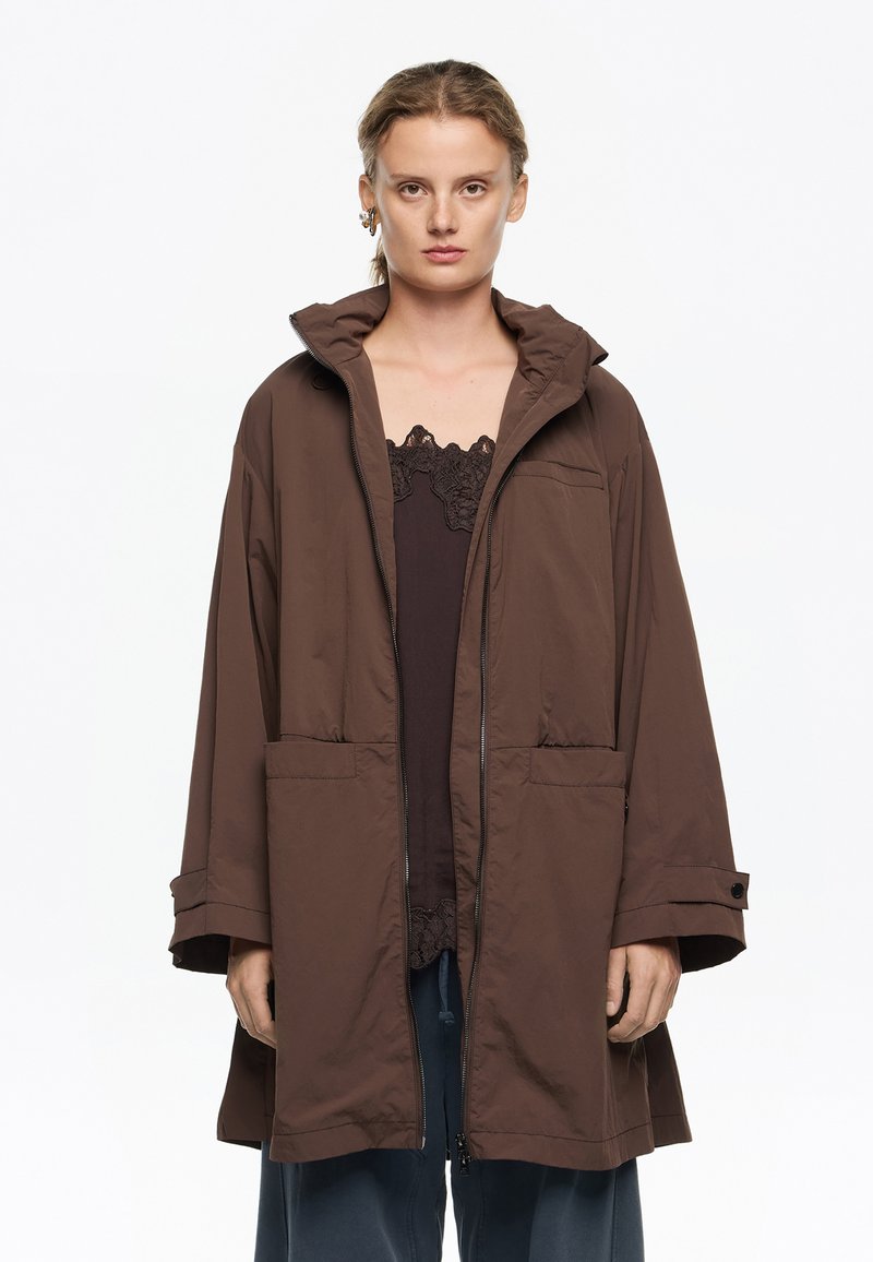 Chaqueta marrón oversized con cremallera y capucha, que cuenta con dos bolsillos delanteros y mangas anchas, hecha de una tela ligera y suave.