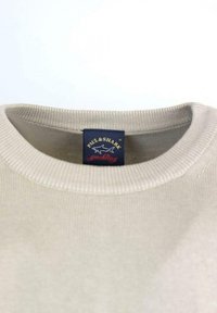 Felpa in cotone beige con colletto a girocollo a coste, con etichetta blu con testo giallo e logo di uno squalo, esposta su uno sfondo bianco.