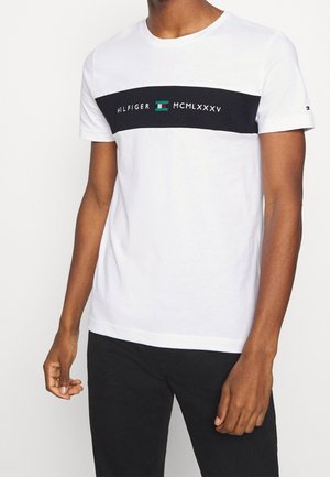 Hvid bomuld T-shirt med en sort horisontal stribe og blå og grøn logo tekst. Kortærmet, rund hals og afslappet pasform.