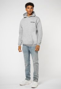 Grijze fleece hoodie met een voorzak en "WORLDWIDE" in het zwart gedrukt. Gedragen met lichtblauwe jeans en witte sneakers.