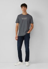 Szary t-shirt z krótkim rękawem z białym napisem "S.OLIVER Elevated Casual", w połączeniu z ciemnymi dżinsami i białymi trampkami.