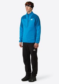 Pullover in pile blu con colletto a mezza zip, pantaloni neri e calzature scure. Le caratteristiche includono logo, pannelli di colore contrastante e una texture liscia.