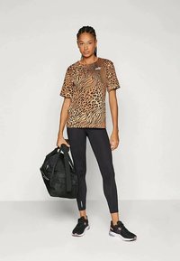 T-shirt de estampado animal castanho e preto com mangas curtas, combinado com leggings pretas e sapatilhas pretas. Uma mala de ginásio preta é segurada com uma das mãos.