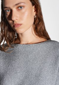 Grijze metallic trui met een brede halslijn, gestructureerde stof en een slim fit. Haar is golvend en schouderlang; oorbel is een kleine ring.
