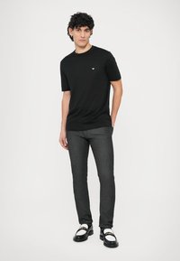 Juoda trumpomis rankovėmis marškinėliai su mažu logotipu ant krūtinės, derinami su tamsiai pilkais siaurais džinsais ir juodai baltais batais.
