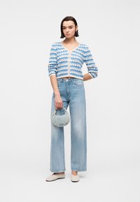 Νεαρή γυναίκα που φοράει μπλε ριγέ ζακέτα, ανοιχτόχρωμα τζιν παντελόνια με φαρδιά πόδια, μπεζ μπαλαρίνες και κρατά μια μικρή τσάντα από τζιν.