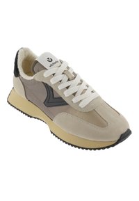 Chaussure de sport beige avec une tige en suède et cuir, lacets blancs, accents noirs et semelle en caoutchouc noir texturé. Étiquette sur la languette.