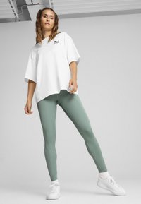 Witte oversized t-shirt met een zwart logo, gecombineerd met nauwsluitende mintgroene leggings en witte sportschoenen. Minimalistisch ontwerp en zachte textuur.