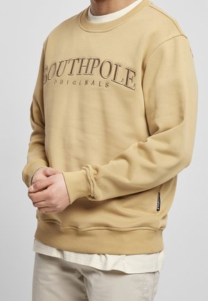 Person wearing a beige sweatshirt with "SOUTHPOLE ORIGINALS" embroidered on the chest, paired with light-colored pants and a white undershirt.