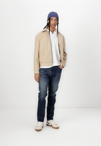 Veste beige au design structuré, chemise blanche à côtes, bonnet bleu, jeans en denim et baskets blanches avec semelles en gomme.