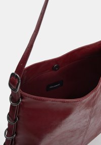 Burgunderfarbene Lederhandtasche mit glatter Textur, ausgestattet mit einem einzigen Tragegurt und Metallringakzenten auf einer Seite. Futter ist schlicht.