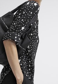 Schwarzes Hemd mit weißem Sternmuster, hochgerollten Ärmeln, kombiniert mit einer schwarzen Tasche, die strukturiertes Material und ein lässiges Design präsentiert.