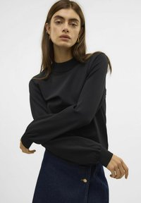Un haut noir à manches longues avec un col rond et des poignets élastiques, associé à une jupe en denim bleu foncé présentant un détail de boutons visible.