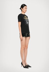 Young woman standing in black graphic t-shirt, shiny black shorts, and black pointed high heels against white background.