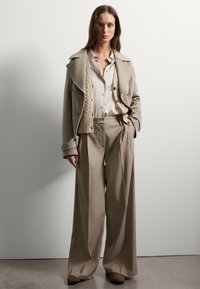 Chaqueta beige de mezcla de lana con botones dobles y amplios solapas, combinada con una blusa de crema clara y pantalones beiges holgados, de pie sobre un suelo gris.