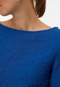 Donna che indossa un top off-shoulder royal blue testurizzato, con i capelli raccolti e la testa leggermente girata di lato.