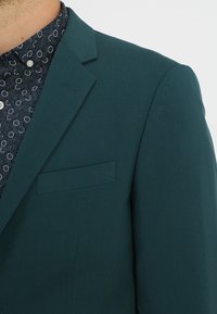 Teal sako s krojem po mjeri, s jednim džepom na prsima i reverima, preko tamno uzorčene košulje s bijelim akcentima.